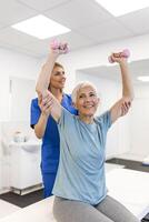 Oder aged woman doing physiotherapy with support from physiotherapists . Senior elderly female sitting in clinic using dumbbells workout exercise for patient with caregiver in nursing care. photo