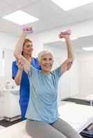 Oder aged woman doing physiotherapy with support from physiotherapists . Senior elderly female sitting in clinic using dumbbells workout exercise for patient with caregiver in nursing care. photo