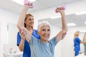 Oder aged woman doing physiotherapy with support from physiotherapists . Senior elderly female sitting in clinic using dumbbells workout exercise for patient with caregiver in nursing care. photo