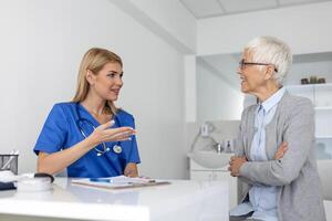 joven mujer médico o gp en blanco médico uniforme consultar mayor hembra paciente en privado hospital. hembra terapeuta hablar hablar con mujer cliente en consulta en clínica. foto