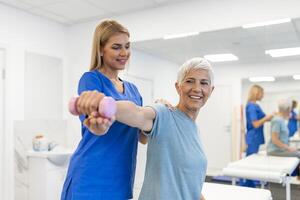 Oder aged woman doing physiotherapy with support from physiotherapists . Senior elderly female sitting in clinic using dumbbells workout exercise for patient with caregiver in nursing care. photo