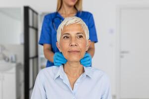 Friendly woman doctor wearing gloves checking sore throat or thyroid glands, touching neck of senior female patient visiting clinic office. Thyroid cancer prevention concept photo