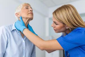 simpático mujer médico vistiendo guantes comprobación dolorido garganta o tiroides glándulas, conmovedor cuello de mayor hembra paciente visitando clínica oficina. tiroides cáncer prevención concepto foto