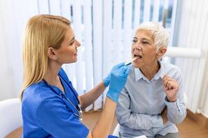 Senior patient opening her mouth for the doctor to look in her throat. Female doctor examining sore throat of patient in clinic. Otolaryngologist examines sore throat of patient. photo