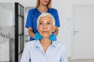 simpático mujer médico vistiendo guantes comprobación dolorido garganta o tiroides glándulas, conmovedor cuello de mayor hembra paciente visitando clínica oficina. tiroides cáncer prevención concepto foto