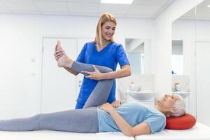 close up of female physiotherapist is helping senior elder woman stretching her hamstring and doing thigh or leg rehabilitation in exercise room - she is lying on massage bed photo