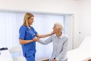 Doctor giving hope. Close up shot of young female physician leaning forward to smiling elderly lady patient holding her hand in palms. Woman caretaker in white coat supporting encouraging old person photo