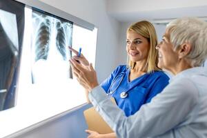 Doctor woman checking x-ray film in medical laboratory at hospital, explaining the results of scan lung on screen to senior patient. photo