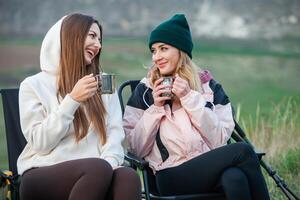 Two beautiful girls sitting in a folding chair and drinking tea in nature. Travel, traveler. photo