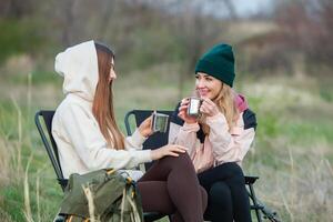 Two beautiful girls sitting in a folding chair and drinking tea in nature. Travel, traveler. photo