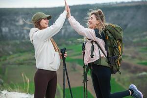 dos joven mujer amigos caminantes con mochilas y trekking polos en parte superior de un montaña. viajar, viajero. foto