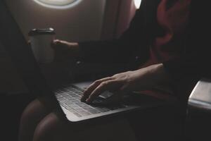 Blonde female tourist checking incoming notification on smartphone sitting on seat of airplane with netbook.Young businesswoman share media from telephone on laptop computer during plane flight photo