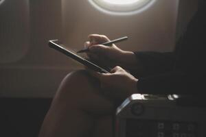 Blonde female tourist checking incoming notification on smartphone sitting on seat of airplane with netbook.Young businesswoman share media from telephone on laptop computer during plane flight photo