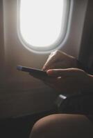 Blonde female tourist checking incoming notification on smartphone sitting on seat of airplane with netbook.Young businesswoman share media from telephone on laptop computer during plane flight photo