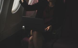 Blonde female tourist checking incoming notification on smartphone sitting on seat of airplane with netbook.Young businesswoman share media from telephone on laptop computer during plane flight photo