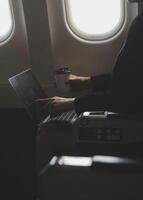 Blonde female tourist checking incoming notification on smartphone sitting on seat of airplane with netbook.Young businesswoman share media from telephone on laptop computer during plane flight photo