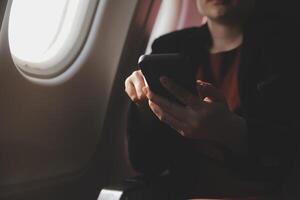 Blonde female tourist checking incoming notification on smartphone sitting on seat of airplane with netbook.Young businesswoman share media from telephone on laptop computer during plane flight photo