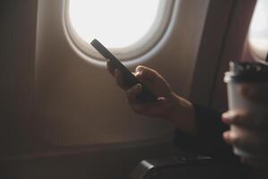 Blonde female tourist checking incoming notification on smartphone sitting on seat of airplane with netbook.Young businesswoman share media from telephone on laptop computer during plane flight photo