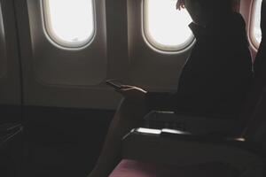 Blonde female tourist checking incoming notification on smartphone sitting on seat of airplane with netbook.Young businesswoman share media from telephone on laptop computer during plane flight photo