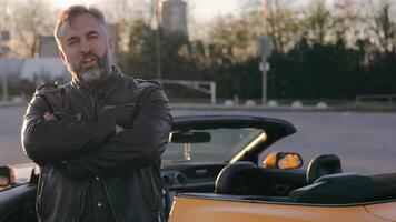 A man in a leather jacket near a convertible. Portrait, A rugged man stands with crossed arms by a convertible at sunset video