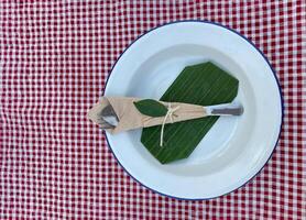 white galvanized plate is paired with a set of spoons and forks tied with hemp rope . Decorate the plate with banana leaves and place it on a red and white checkered tablecloth. photo