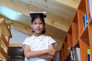 Asian girl standing with her arms crossed in front of a bookshelf and books on her head. Concept child research, education, development, brain, student, knowledge, learning. photo