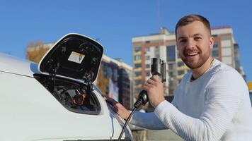 A light-skinned male driver holds a charging cable for an electric car sitting near an open car battery and blows out imaginary smoke from the charging cable, as if after a shot video