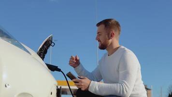 A fair-skinned male driver connects an electric car to the power system to charge the car battery and controls the charging process via his smartphone video