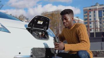 Dark-skinned male driver connects electric car to power system to charge car battery video