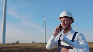 A male engineer in a white helmet, work blue overalls and a classic shirt holds in his hand a large rolled sheet of paper with drawings of the project and talks on the cellphone near the wind turbines video