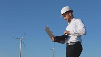 A male engineer in a white helmet and a classic shirt inspects the power plant's capacity and makes notes in a laptop video