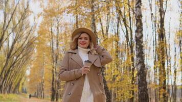 giovane donna nel un' Marrone cappotto e cappello passeggiate nel il autunno parco e bevande caffè video