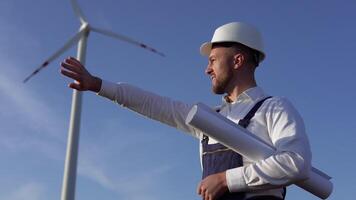 un masculino ingeniero en un blanco casco, trabajo azul mono y un clásico camisa examina el viento turbinas de un poder planta video