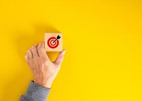 Hand holding wooden block with dartboard with arrow on yellow background photo