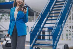 joven asiático mujer en internacional aeropuerto, utilizando móvil teléfono inteligente y comprobación vuelo a el vuelo información tablero foto