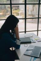 mujeres asiáticas sentadas en una oficina en casa con estrés y tensión ocular. mujer de negocios cansada sosteniendo anteojos y masajeando el puente nasal. hay tabletas, computadoras portátiles y café. foto