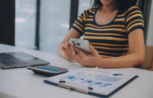 encantadora joven mujer de negocios asiática sentada en una computadora portátil en la oficina, haciendo un informe que calcula el documento de verificación del servicio de ingresos internos del saldo. foto