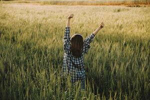 Young pretty woman in red summer dress and straw hat walking on yellow farm field with ripe golden wheat enjoying warm evening. photo