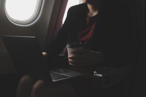 Blonde female tourist checking incoming notification on smartphone sitting on seat of airplane with netbook.Young businesswoman share media from telephone on laptop computer during plane flight photo