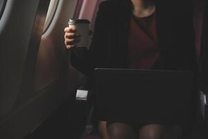 Blonde female tourist checking incoming notification on smartphone sitting on seat of airplane with netbook.Young businesswoman share media from telephone on laptop computer during plane flight photo