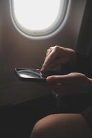 Blonde female tourist checking incoming notification on smartphone sitting on seat of airplane with netbook.Young businesswoman share media from telephone on laptop computer during plane flight photo