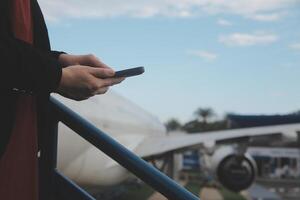 joven asiático mujer en internacional aeropuerto, utilizando móvil teléfono inteligente y comprobación vuelo a el vuelo información tablero foto