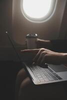 Blonde female tourist checking incoming notification on smartphone sitting on seat of airplane with netbook.Young businesswoman share media from telephone on laptop computer during plane flight photo