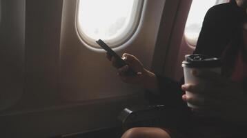 Blonde female tourist checking incoming notification on smartphone sitting on seat of airplane with netbook.Young businesswoman share media from telephone on laptop computer during plane flight photo