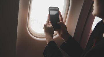 Blonde female tourist checking incoming notification on smartphone sitting on seat of airplane with netbook.Young businesswoman share media from telephone on laptop computer during plane flight photo