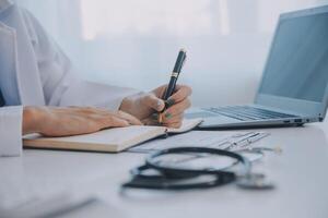 Doctor and patient sitting at the desk in clinic office. The focus is on female physician's hands filling up the medication history record form, close up. Medicine concept photo