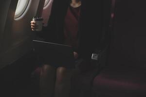Blonde female tourist checking incoming notification on smartphone sitting on seat of airplane with netbook.Young businesswoman share media from telephone on laptop computer during plane flight photo