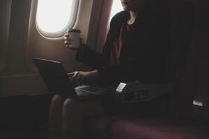 Blonde female tourist checking incoming notification on smartphone sitting on seat of airplane with netbook.Young businesswoman share media from telephone on laptop computer during plane flight photo