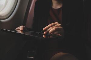 Blonde female tourist checking incoming notification on smartphone sitting on seat of airplane with netbook.Young businesswoman share media from telephone on laptop computer during plane flight photo