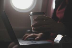 Blonde female tourist checking incoming notification on smartphone sitting on seat of airplane with netbook.Young businesswoman share media from telephone on laptop computer during plane flight photo